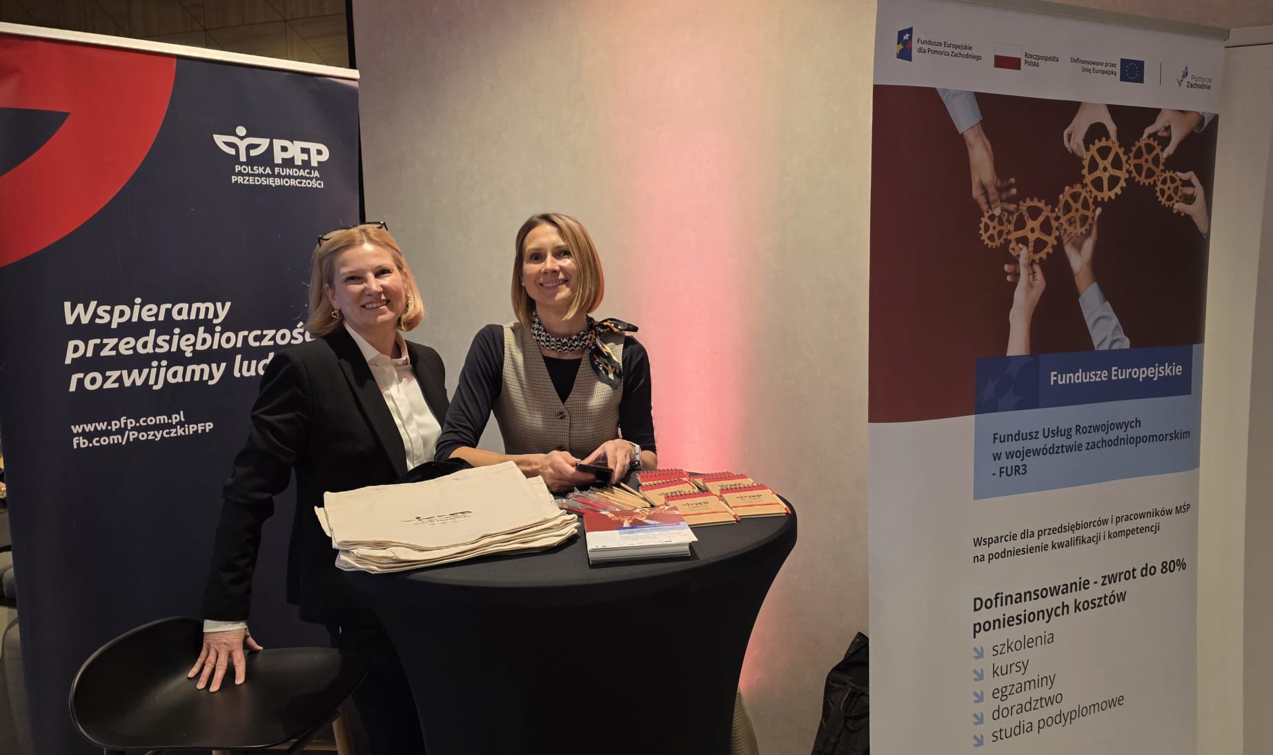 Dwie uśmiechnięte kobiety (przedstawicielki PFP) stoją za stoiskiem informacyjnym PFP, na którym widoczne są materiały promocyjne i ulotki. W tle znajduje się baner promujący 'Fundusze Usług Rozwojowych w województwie zachodniopomorskim - FUR3' oraz PFP.